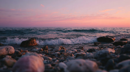 An image captures a coastal scene during sunset, showcasing the interaction of ocean waves and a pebbled shoreline. The colors are dominated by hues of pink and orange in the sky, reflecting on the water. The composition is a low-angle shot, providing a close-up view of the rocks and waves. This could be used for various editorial or commercial purposes.の素材