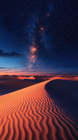 A striking desert landscape features textured sand dunes leading towards the horizon. The night sky presents a dark blue backdrop illuminated by countless stars and the Milky Way. The composition is lit by warm, reddish hues, suggesting a sunset or sunrise, and could be utilized for various commercial projects.の素材