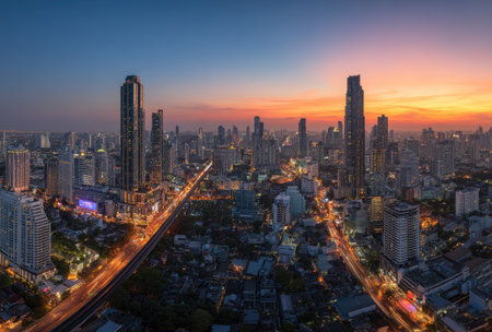 An aerial view presents a sprawling city skyline at sunset. Modern skyscrapers and buildings dominate the frame, bathed in the warm glow of the setting sun. The composition highlights contrasting colors and textures, capturing a dynamic urban environment. Suitable for commercial projects or editorial use.の素材