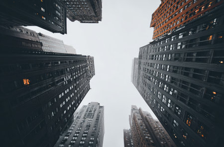 An upward perspective captures the imposing height of multiple skyscrapers against a subdued, cloudy sky. The buildings display various architectural styles and muted color palettes. This cityscape scene, with its strong lines and depth, could serve in diverse editorial and commercial contexts.の素材
