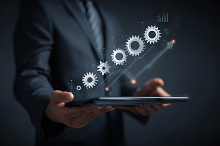 A person in a suit holds a digital tablet, with projected gear icons and a growing graph overlaying it. The image uses a dark color palette. The composition has shallow depth of field, with soft lighting enhancing the central focus. Suitable for business, finance, and technology applications.の素材
