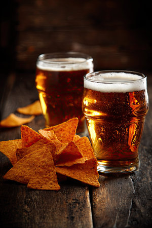 Two glasses of amber-colored beer are displayed with a pile of tortilla chips. The image showcases a rustic aesthetic with warm lighting, highlighting textures. This setup suggests a casual gathering. Suitable for advertising, culinary, or social media content.の素材