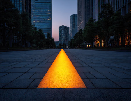 An illuminated pathway glows brightly through a city at dusk. The scene showcases a geometric composition, with an orange-yellow light contrasting against the dark paving and buildings. The lighting creates a dramatic atmosphere. Suitable for a range of commercial applications, this image is visually captivating.の素材