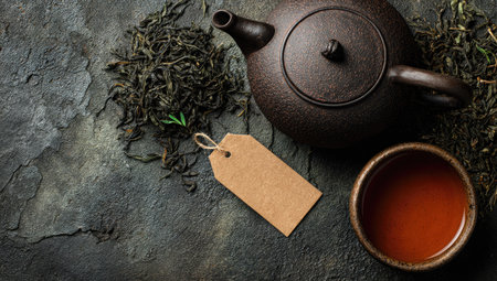 An overhead shot presents a brown teapot, a filled teacup, and dried tea leaves scattered on a textured, dark surface. The image showcases natural lighting, with a focus on color and texture. Suitable for diverse editorial and commercial projects, this composition is ideal for illustrating tea-related themes.の素材