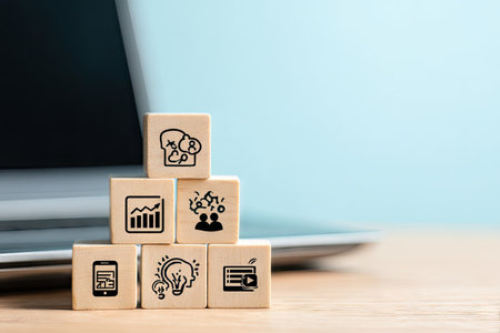 A pyramid of wooden blocks displaying business-related icons sits in front of a laptop. The composition features a shallow depth of field, with soft lighting and neutral tones. It suggests concepts of technology, strategy, and business solutions. The image could be used for various commercial or editorial purposes.の素材