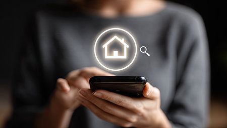 A person holds a smartphone, displaying a glowing house icon within a circle. The image showcases a modern, tech-focused composition. The person's blurred form and neutral tones contrast with the bright symbol. This scene evokes the concept of property search, suggesting potential commercial applications.の素材