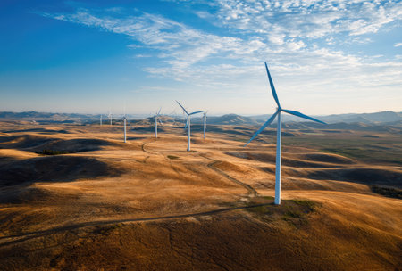 The image features several wind turbines positioned across a field. The scene showcases a sunny day with a clear blue sky and rolling hills. The color palette primarily includes shades of brown, green and blue. This composition is suitable for illustrating concepts of renewable energy and sustainability.の素材