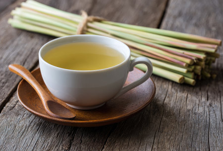 A close-up captures a cup of herbal tea, placed on a saucer with a wooden spoon. Fresh lemongrass stalks are arranged nearby. The image features a natural, warm color palette, and is lit by soft light. Ideal for projects related to wellness, food, and beverages.の素材