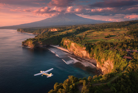 An airplane soars near a scenic coastline, with a majestic mountain in the distance. The image showcases the natural beauty of the area with lush greenery and a sandy beach. The composition captures the vibrant colors of sunset, offering a visually appealing scene suitable for various commercial uses.の素材