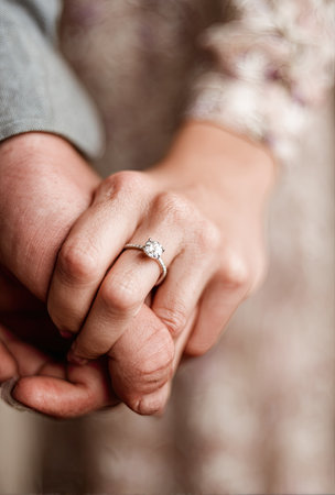 This image showcases hands clasped together, with a prominent engagement ring on one finger. The soft lighting highlights the texture of the skin and the intricate details of the ring. The overall composition suggests a moment of connection and promise. Suitable for various editorial and commercial applications.の素材