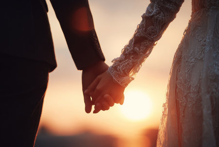 A couple holds hands with the sunset in the background during a wedding ceremony. The image displays a close-up of the hands, the woman wearing a lace-sleeved dress. Warm tones dominate the scene, suggesting an outdoor setting at dusk. Suitable for editorial or commercial purposes.の素材