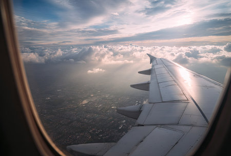An airplane wing extends into a cloudy sky visible from a window. The scene features varying shades of blue, white and gray. The image showcases natural light, possibly indicating daytime. This composition can be used for travel, transportation or aviation related materials.の素材
