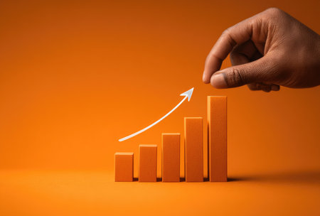 A dark-skinned hand places a block on a rising bar graph, with an upward-pointing arrow. The image features a solid orange background and a simple, graphical style. It could be used in business presentations, financial reports, or marketing materials to visually represent growth or progress.の素材
