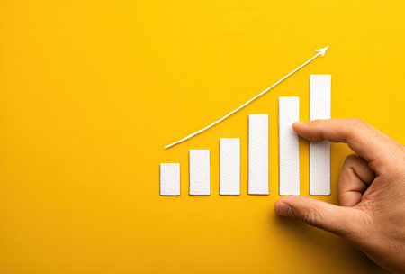 A hand interacts with a paper bar graph against a vibrant yellow backdrop. The composition features a rising white line symbolizing growth. The scene uses a flat, overhead perspective. This imagery could be employed for presentations on business, finance, or economic progress.の素材