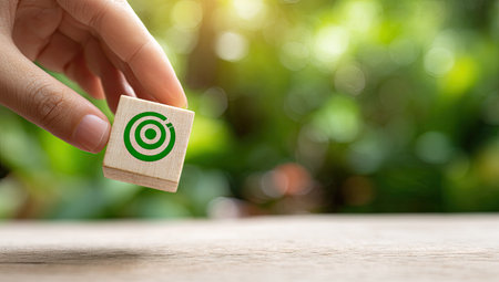 A hand delicately holds a small wooden block featuring a green target symbol. The image showcases a shallow depth of field, with a vibrant green backdrop suggesting an outdoor environment. The composition conveys concepts of precision, goals, and focus. Suitable for business and marketing visuals.の素材