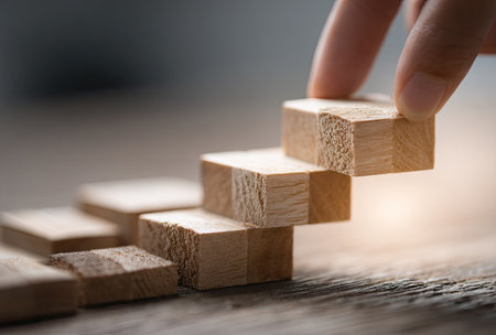 A hand places a wooden block onto a structure resembling stairs. The warm-toned image features wooden blocks on a wooden surface. The lighting suggests an indoor setting, possibly during the daytime. This image is suitable for various commercial uses, including illustrating concepts related to business or personal development.の素材
