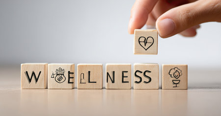 A hand places a block with a heart icon onto a set of blocks spelling "WELLNESS". The image features a shallow depth of field, with soft lighting and a neutral background. This visual could be used in various commercial projects highlighting health, well-being, or lifestyle topics.の素材
