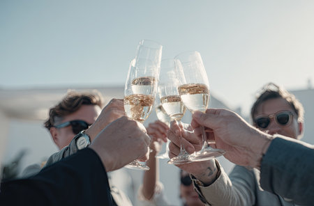 Several people raise champagne glasses in a celebratory toast, likely during an outdoor gathering. The image showcases the clear glasses filled with a light-colored liquid against a soft, blurred background. The composition suggests a moment of joy, potentially suitable for editorial content or promotional materials.の素材