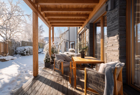 An inviting outdoor scene shows a wooden deck with seating under a wooden overhang. The deck overlooks a snow-covered landscape. Natural light illuminates the scene, highlighting the textures of wood and fabrics. This image can be used for editorial purposes or to illustrate themes of outdoor living.の素材