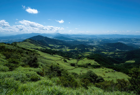 An expansive view showcases rolling hills covered in lush green vegetation under a vast blue sky dotted with clouds. The composition features a natural landscape with variations in tone and texture. This image could be used for various commercial and editorial applications, providing a sense of natural beauty and tranquility.の素材