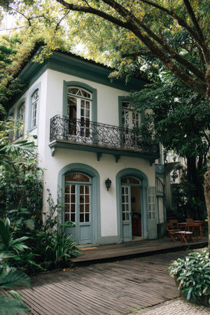 A classic two-story house, painted white with blue-green accents, stands amidst abundant greenery. The building features ornate balconies and windows. Natural light and composition create a visually appealing facade. Suitable for architectural, residential, and lifestyle commercial applications.の素材