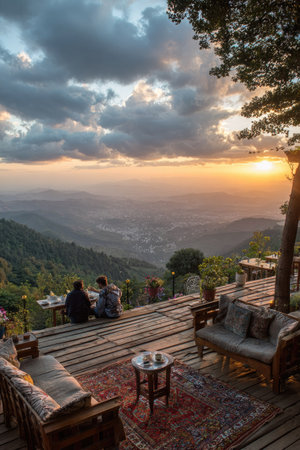 Two individuals sit on a deck overlooking a mountain range as the sun sets. The scene features a natural environment with vibrant colors. The composition utilizes a wide angle and warm lighting. The image may be suitable for travel, lifestyle, or environmental projects.の素材