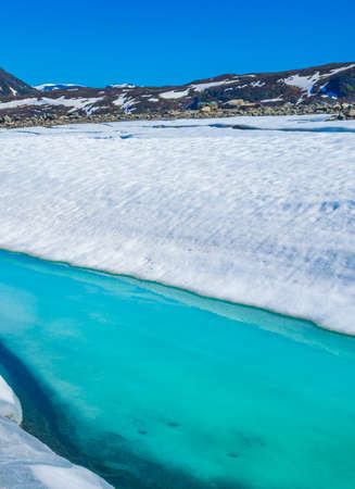 Frozen turquoise lake Vavatn panorama in summer landscape and mountains with snow in Hemsedal...