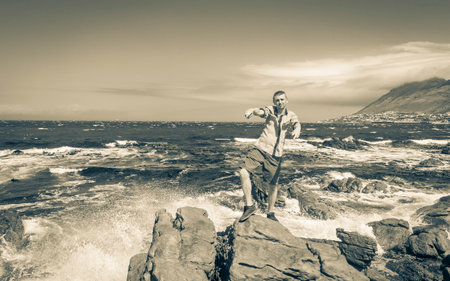 Male model tourist traveler at False Bay rough coast landscape with boulders waves and mountains with clouds in Glencairn Simons Town Cape Town Western Cape South Africa.の写真素材