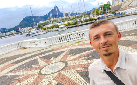 Handsome man tourist takes selfie in the Urca Village coastal and beach promenade with street sidewalk pier harbor jetty yachts and boats in Rio de Janeiro Brazil.の写真素材