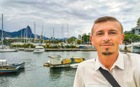 Handsome man tourist takes selfie in the Urca Village coastal and beach promenade with street sidewalk pier harbor jetty yachts and boats in Rio de Janeiro Brazil.の写真素材
