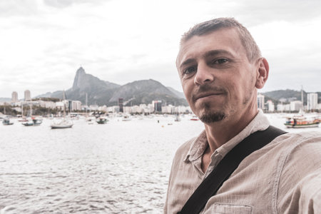 Handsome man tourist takes selfie in the Urca Village coastal and beach promenade with street sidewalk pier harbor jetty yachts and boats in Rio de Janeiro Brazil.の写真素材