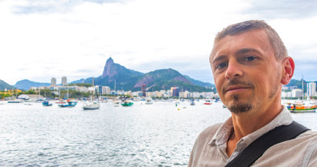 Handsome man tourist takes selfie in the Urca Village coastal and beach promenade with street sidewalk pier harbor jetty yachts and boats in Rio de Janeiro Brazil.の写真素材