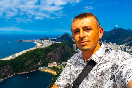 Handsome Man Tourist with the Rio de Janeiro Panorama View of the City Skyline Beaches Beach Coast Sea Mountains Tropical Forest and Blue Cloudy Sky in State of Rio de Janeiro Brazil.の写真素材