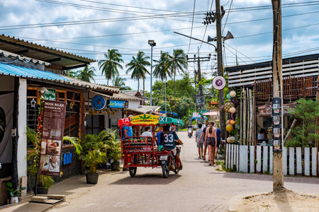 Koh Mook, TH - 23.12.2024: Tourists and locals exploring a vibrant village street lined with shops, restaurants, and palm trees, with motorcycle taxis providing transportation in Kantang, Trang,のeditorial素材