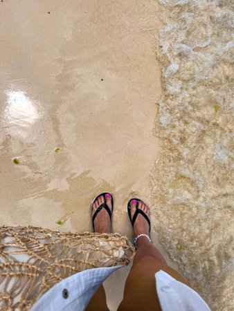 Top view of female feet in slippers standing on yellow sand with a sea wave