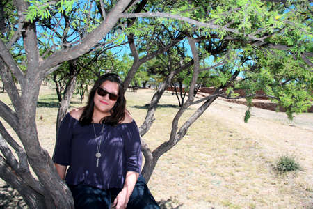 Latin adult woman with sunglasses sitting on a tree looks her pensive and relaxed face among the branches calm, happy and in harmonyの写真素材