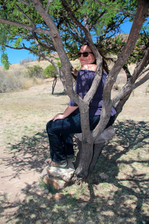 Latin adult woman with sunglasses sitting on a tree looks her pensive and relaxed face among the branches calm, happy and in harmonyの写真素材