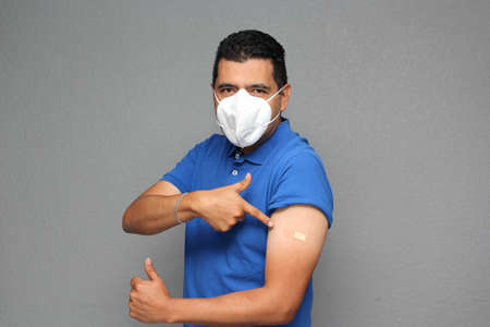 Happy Latino adult man shows his arm that just received the  vaccine and has an adhesive band in the...