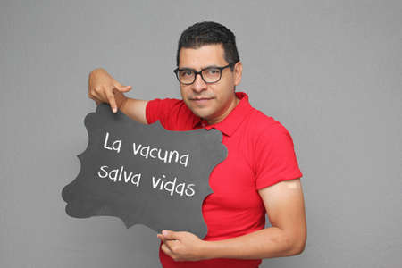 Latino adult man shows his arm that just received the  vaccine and a sign that says The vaccine...