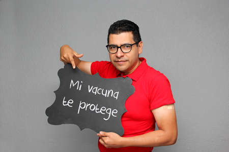 Latino adult man shows his arm that just received the  vaccine and a sign that says My vaccine...