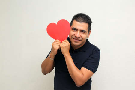 Latin adult man shows a big red heart for his enthusiasm for the arrival of February and celebrate Valentine's Day of Love and Friendship with his family, friends and partner with happiness and affectの写真素材