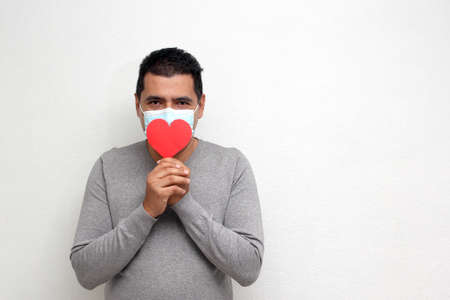 Latino adult man with face mask as protection in the new normality due to the Covid-19 pandemic shows a red heart to celebrate Valentine's Day of Love and Friendshipの写真素材