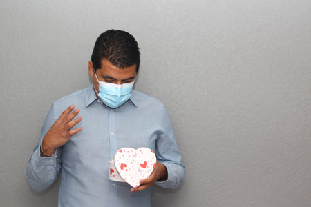 Valentine's new normal. Latin man with blue shirt, clinical use protection mask and heart box of chocolatesの写真素材
