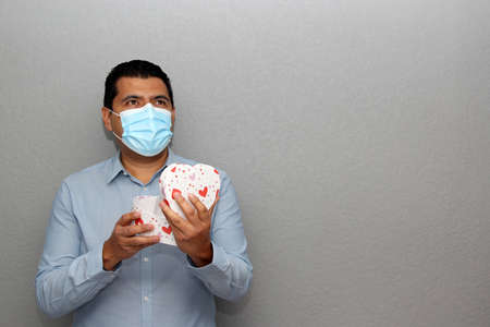 Valentine's new normal. Latin man with blue shirt, clinical use protection mask and heart box of...