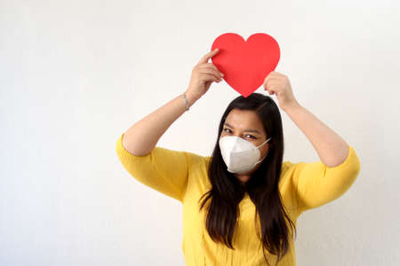 Latin adult woman with a yellow blouse and a big red heart shows her enthusiasm for Valentine's Day of Love and Friendship with a face mask in the new normality due to the pandemicの写真素材