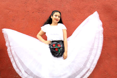Young Latin Mexican woman is happy because she is wearing a regional white dress from Veracruz Mexico modeling the dance steps in front of a red wallの写真素材