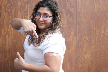Latin adult woman with white blouse wavy hair and glasses shows her arm with a bandage because she...