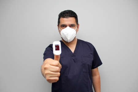 Latin adult man with medical uniform and mask shows a pulse oximeter in his hand to measure...