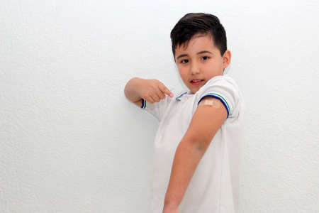 8-year-old Hispanic boy wearing a school uniform shirt happily shows his recently vaccinated arm...