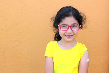 Happy little Hispanic girl with wavy hair wearing a yellow t-shirt and red glasses shows her recently vaccinated arm against Covid-19 in the new normality due to the Coronavirus pandemicの写真素材